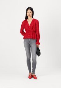 Blusa rossa, con texture, maniche lunghe e design avvolgente; abbinata a jeans skinny grigi e ballerine rosse; tiene una borsa nera.