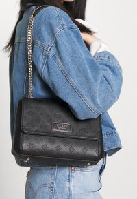 Zwarte schoudertas van imitatieleer met een reliëflogo patroon, zilveren kettingriem en magnetische klepsluiting. Moderne rechthoekige vorm.