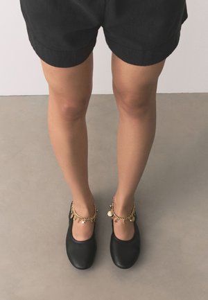 Jambes portant un short noir et des chaussures plates noires avec des bracelets de cheville à breloques dorées sur un sol gris et un mur blanc en arrière-plan.