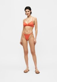 Γυναίκα όρθια, φοράει πορτοκαλί bralette και αντιστοιχισμένα σλιπ ψηλού κόστους με μπεζ σανδάλια χωρίς κούμπωμα σε απλό λευκό φόντο.