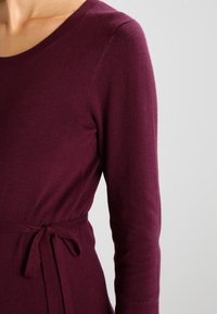 Bordowy top z długim rękawem i okrągłym dekoltem, z wiązaniem w talii. Miękki materiał, dopasowany krój i gładka tekstura.