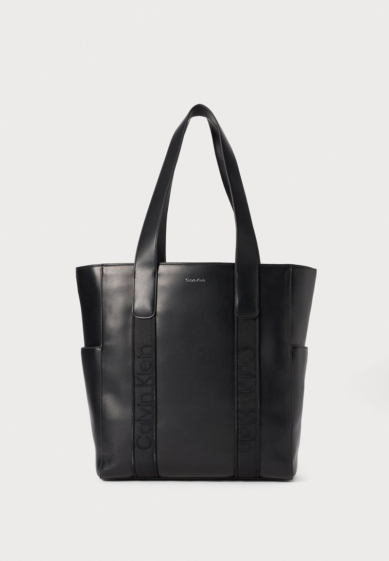Sac en cuir noir avec double anses, présentant des détails de logo en relief et des poches latérales pour un espace de rangement supplémentaire. Fini lisse, design structuré.