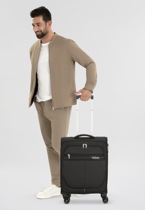 Valise noire à coque rigide, deux poches avant zippées, quincaillerie en argent et quatre roues. Un homme en tenue beige tient la valise.