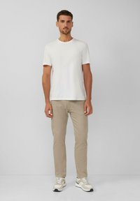 Man die tegen een eenvoudige muur staat, gekleed in een wit T-shirt, beige broek en beige en witte sneakers.