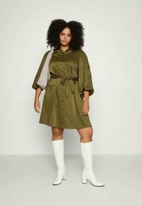 Vestido camiseiro verde oliva com cinto, com botões na frente, mangas oversized e tecido suave; combinado com botas brancas até ao joelho.