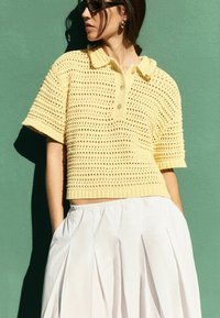 Mujer con gafas de sol y pendientes dorados, vestida con un top amarillo de crochet de manga corta y una falda blanca plisada, de pie frente a una pared verde.