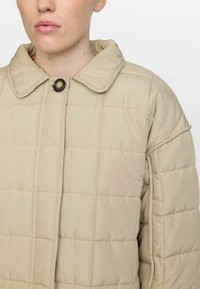 Beige, kviltet pufferjakke med en struktureret krave, frontknaplukning og synlig syningsmønster. Glat, letvægtsstof.