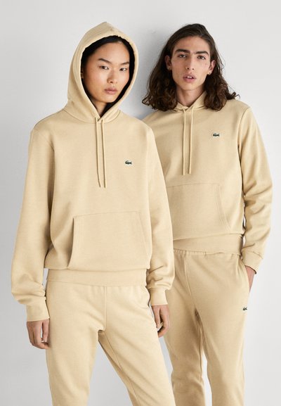 Ensemble de hoodie et de sweatpants beige, avec une poche frontale, une capuche à cordon de serrage et des accents de logo verts. Tissu doux avec un design décontracté.