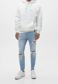 Bílá mikina s kapucí a přední kapsou, světle modré roztrhané džíny a bílé tenisky s hnědými detaily, zobrazené na jednoduchém pozadí.