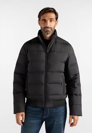 Uomo con barba e capelli corti che indossa una giacca imbottita nera e jeans blu, in piedi davanti a uno sfondo chiaro e uniforme.