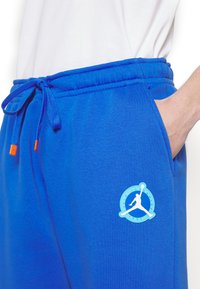Personne portant un pantalon de survêtement bleu vif avec un cordon et un logo de joueur de basket-ball près de la poche, main partiellement dans la poche.
