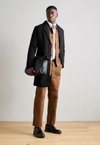 Man standing indoors wearing brown pants, matching jacket, black overcoat, black shoes, holding a black leather briefcase.