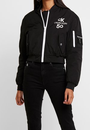 Kvinde iført en sort Calvin Klein bomberjakke med hvid lynlås og logo, parret med sorte højtaljede jeans.