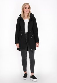 Schwarze wasserdichte Jacke mit Kapuze, tailliertem Schnitt, Reißverschluss vorne und verstellbaren Zugbändern. Kombiniert mit grauen Leggings und schwarzen Turnschuhen.
