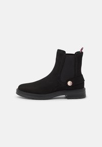 Botas de tobillo de ante negro con paneles laterales elásticos, punta redondeada y un pequeño botón metálico decorativo. Suela de goma plana.