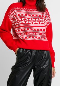 Red knit turtleneck sweater featuring a white holiday pattern, including text that reads "NOËL JOYEUX," with loose fit and ribbed cuffs.