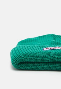 Bonnet en tricot vert avec une texture côtelée. Le bord replié présente un écusson rectangulaire avec le nom de la marque.