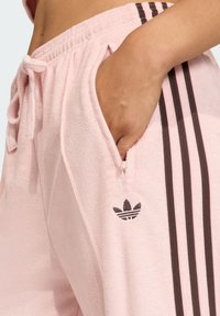 Pink terry cloth joggers feature a drawstring waist, side pockets, and three dark brown stripes along the side with a small logo.