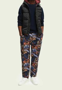 Zwarte gewatteerde vest over een blauwe trui, gecombineerd met een broek met een patroon in blauw, bruin en groen. Witte sneakers maken de look compleet.