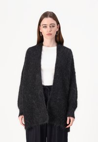 Svart, fluffy cardigan laget av mykt materiale, med åpen front, vide ermer og en avslappet passform. Båret over en hvit skjorte.
