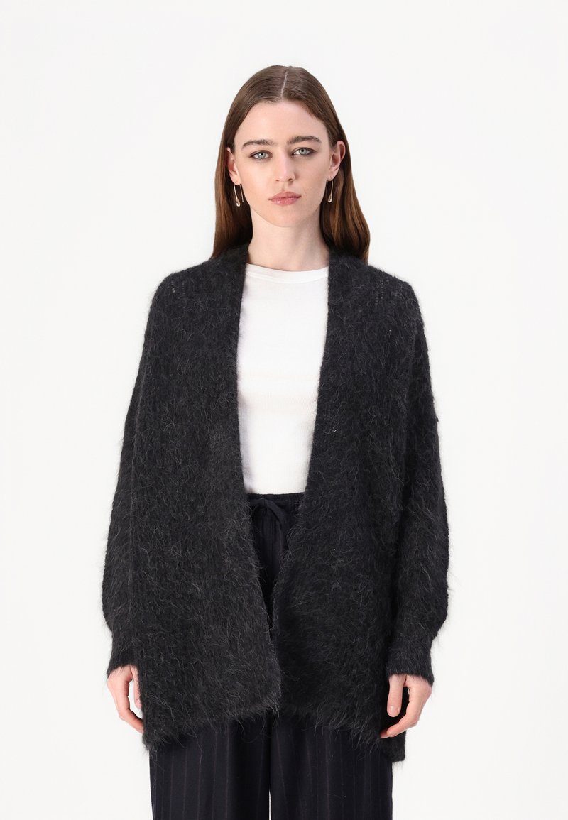 Svart, fluffy cardigan laget av mykt materiale, med åpen front, vide ermer og en avslappet passform. Båret over en hvit skjorte.