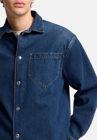 Camicia in denim blu scuro, con colletto a punta, chiusura con bottoni frontali e tasca sul petto. Tessuto strutturato con cuciture a contrasto.