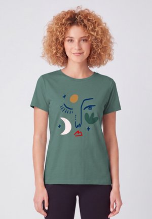 Junge Frau mit lockigem Haar trägt ein grünes T-Shirt mit einem bunten abstrakten Gesicht mit Mond- und Sternformen.
