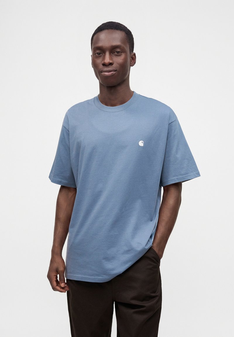 Uomo di colore in piedi con una mano in tasca, indossa una t-shirt celeste a maniche corte e pantaloni scuri su uno sfondo bianco uniforme.