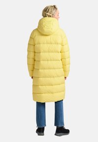 Geel pufferjack met horizontaal gestikt ontwerp, vastgemaakt capuchon en knielange snit. Gecombineerd met blauwe jeans en zwarte schoenen.