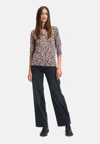 Blumenmuster-Bluse mit langen Ärmeln in Lila, Pink und Beige auf schwarzem Hintergrund, kombiniert mit dunklen, weiten Jeans und Stiefeletten.