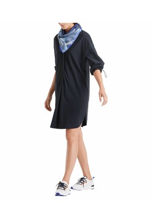 Person geht und trägt ein locker sitzendes schwarzes Kleid mit hochgekrempelten Ärmeln, einen blau gemusterten Schal und weiße Sportschuhe mit schwarzen Schnürsenkeln.
