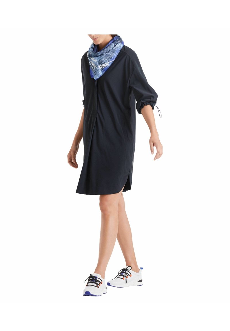 Personne marchant portant une robe noire ample avec des manches retroussées, une écharpe bleue à motifs et des baskets blanches à lacets noirs.