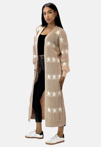 Cárdigan largo de punto beige con patrones de estrellas blancas, de frente abierto y con puños acanalados. Combinado con jeans negros y zapatillas blancas.