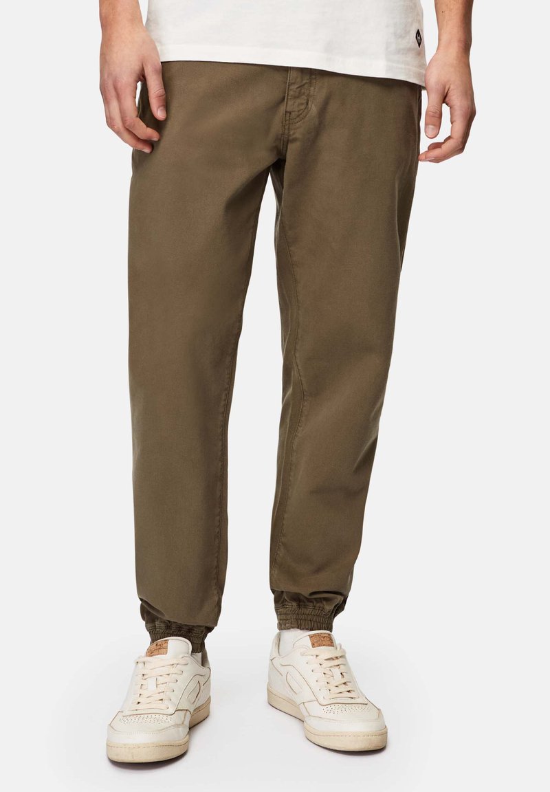 Olivgrüne Chinos mit elastischen Bündchen, die eine glatte Textur und seitliche Taschen aufweisen, kombiniert mit weißen Sneakers.