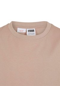 Bēšas krāsas T-krekls ar apaļu kakla izgriezumu, kurš rāda divas iekšējās kakla etiķetes – vienā ir zīmola "Urban Classics" un izmēra informācija, otrā brīdinājums par ugunsdrošību.