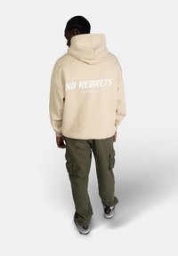 Felpa beige con cappuccio con la scritta "NO REGRETS" in bianco sul retro, abbinata a pantaloni cargo verdi oliva e sneaker.