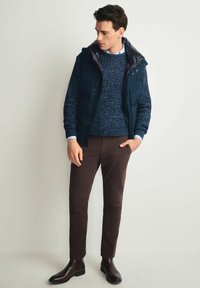 Granatowa kamizelka puchowa z wysokim kołnierzem na granatowym i białym swetrze robionym na drutach, w zestawie z brązowymi spodniami i czarnymi botkami za kostkę. Gładkie tekstury tkanin.