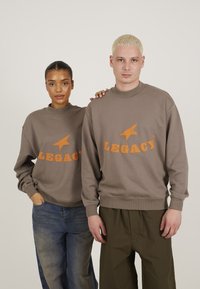 Pulovere cu guler rotund de culoare maro, cu text portocaliu "LEGACY" și un grafic cu stea. Prezentate de două persoane, asortate cu blugi și pantaloni de culoare verde măsliniu.