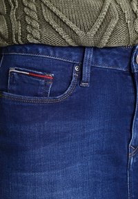 Mörkblå denimjeans med en subtil blekning, som har en liten röd, vit och blå accent på fickan och en standard framknäppning.