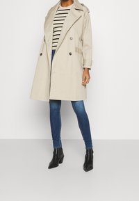 Mujer vestida con un abrigo trench beige, camiseta de rayas, jeans skinny azules y botines negros de punta contra un fondo blanco liso.