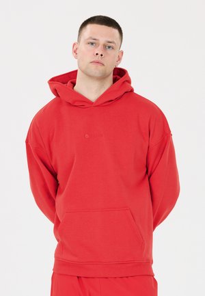 Young man with short hair wearing a red hoodie and matching red pants, standing with hands behind his back against a plain background.