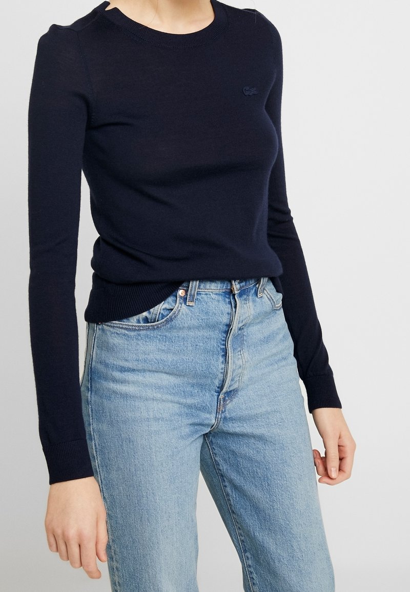 Personne portant un pull ajusté bleu marine à manches longues et un jean taille haute bleu clair sur un fond neutre.