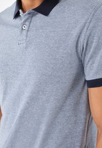 Man som bär en ljusblå kortärmad polo-skjorta med mörkblå krage och ärmslut, visar överkroppen och den vikta kragen.