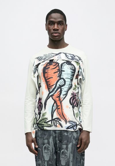 Μπλούζα με μακριά μανίκια σε ανοιχτό κρεμ χρώμα με πολύχρωμο σχέδιο καρότου και λουλουδάτες λεπτομέρειες· φορεμένη με παντελόνι σε σκουρό γκρι αποχρώσεις με μοτίβα.