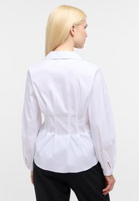 Camicetta bianca aderente con maniche lunghe, dettaglio plissettato sul retro e colletto classico. Texture liscia e forma strutturata.