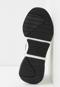 Semelle de chaussure noire avec une bande de roulement à motifs, présentant un logo Puma central dans une section rectangulaire en creux avec des accents blancs et petits jaunes.