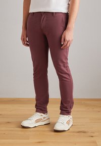 Pantalon slim bordeaux en tissu stretch, avec une coupe classique cinq poches et des détails cousus, associé à des baskets blanches.
