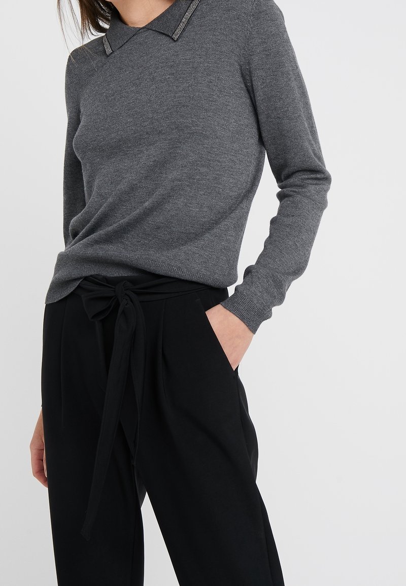 Femme portant un pull gris à manches longues avec un col noir et un pantalon noir taille haute noué avec une ceinture en tissu, main dans la poche.