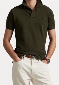 Man i mörkgrön polo med ståkrage, vita byxor, brun läderbälte och brunt armbandsur, med händerna i fickorna.