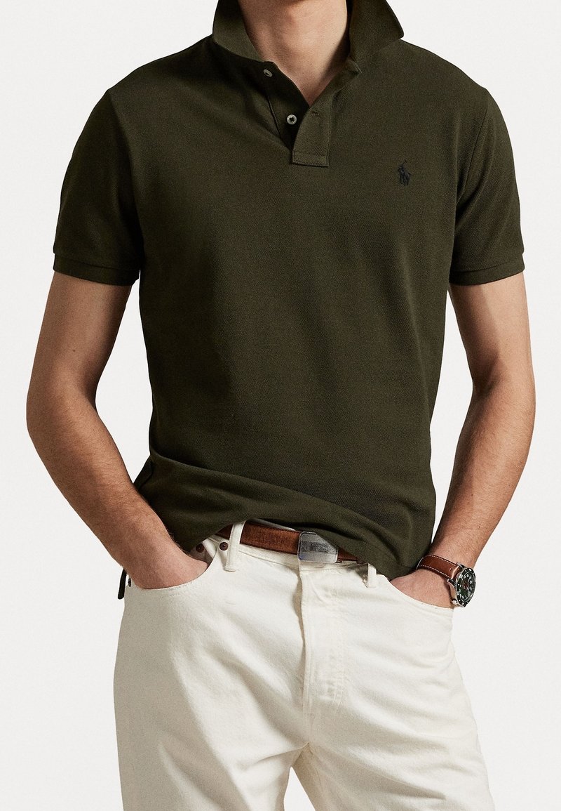 Man i mörkgrön polo med ståkrage, vita byxor, brun läderbälte och brunt armbandsur, med händerna i fickorna.